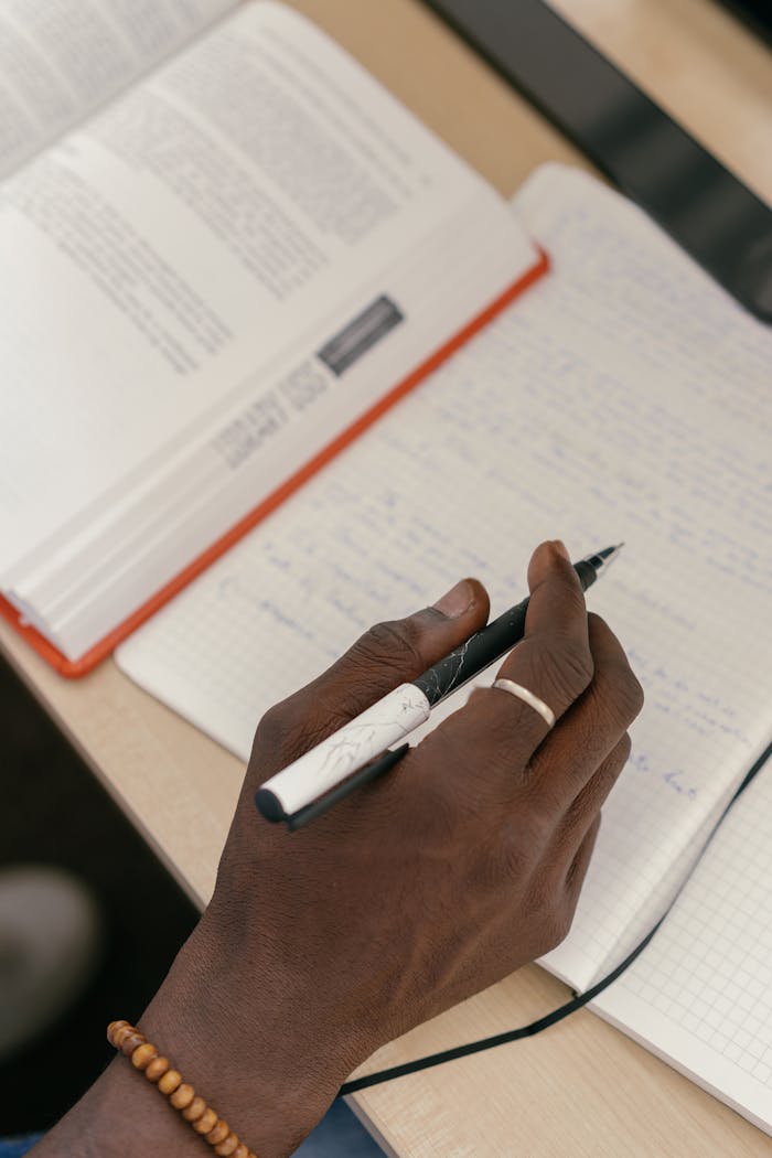 A close-up shot of a hand writing in a notebook with an open book nearby, capturing a study moment.